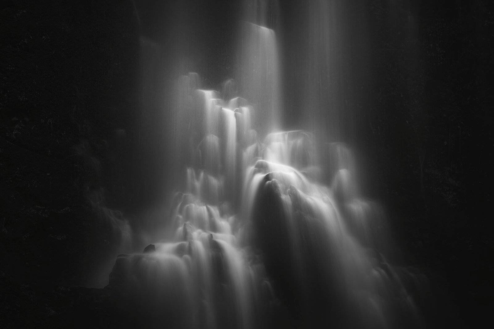 Long-exposure black-and-white image of a multi-tiered waterfall cascading down rocks in a dark forest.