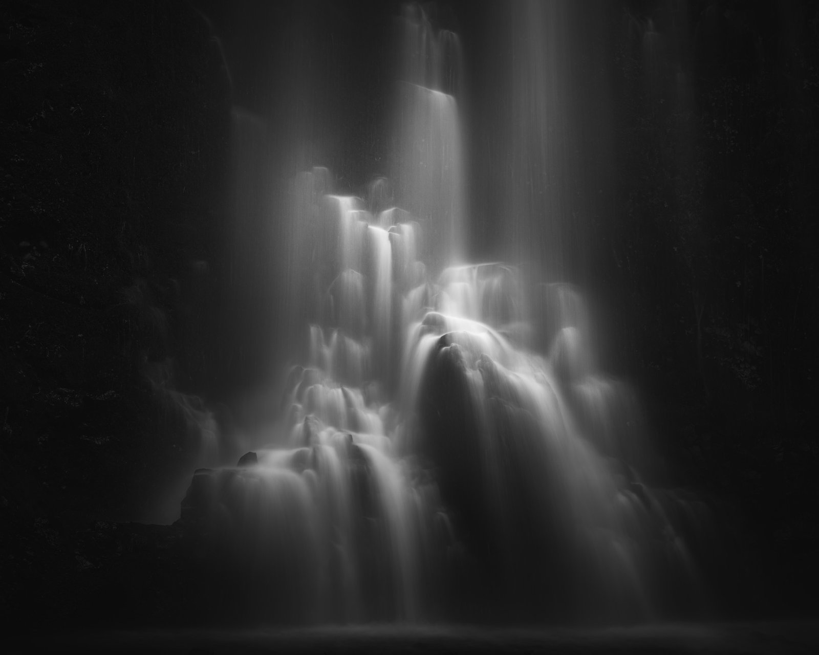 Monochrome image of a multi-tiered waterfall pouring over rocks in a dark, moody landscape with long-exposure blur on the water surface.