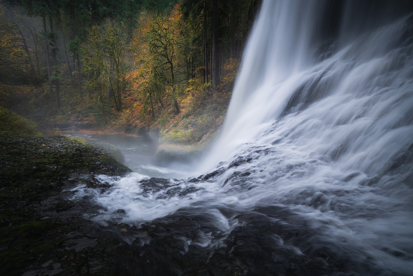 Waterfall cascading down rocky slope on the right, mist at the base, with a dense forest of autumn-colored trees on the left.