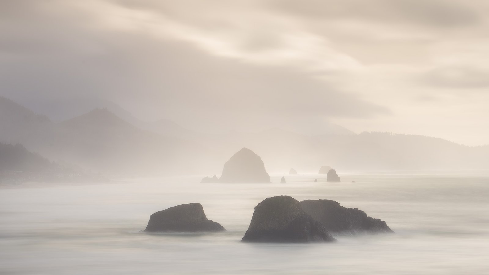 Mist-blanketed coastal scene with calm water and jagged rocks protruding from the sea, distant hills shrouded in fog at sunset.