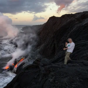 A photographer on rugged lava rocks filming lava entering the sea as steam rises from the water.